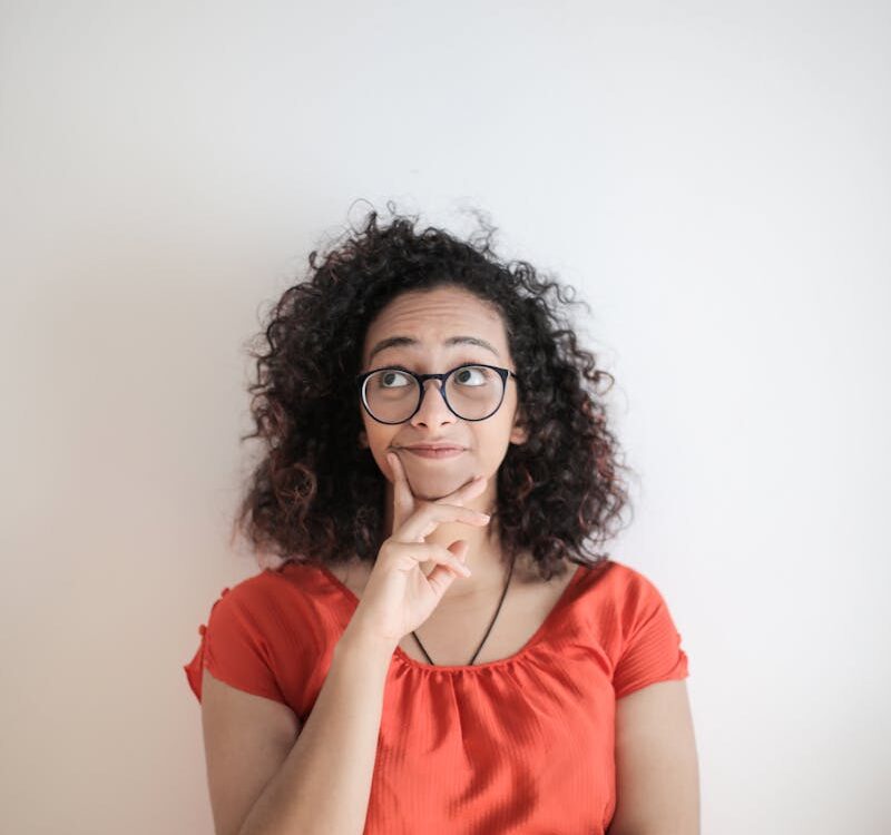 Portrait Photo of Woman in Red Top Wearing Black Framed Eyeglasses Standing In Front of White Background Thinking