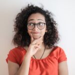 Portrait Photo of Woman in Red Top Wearing Black Framed Eyeglasses Standing In Front of White Background Thinking